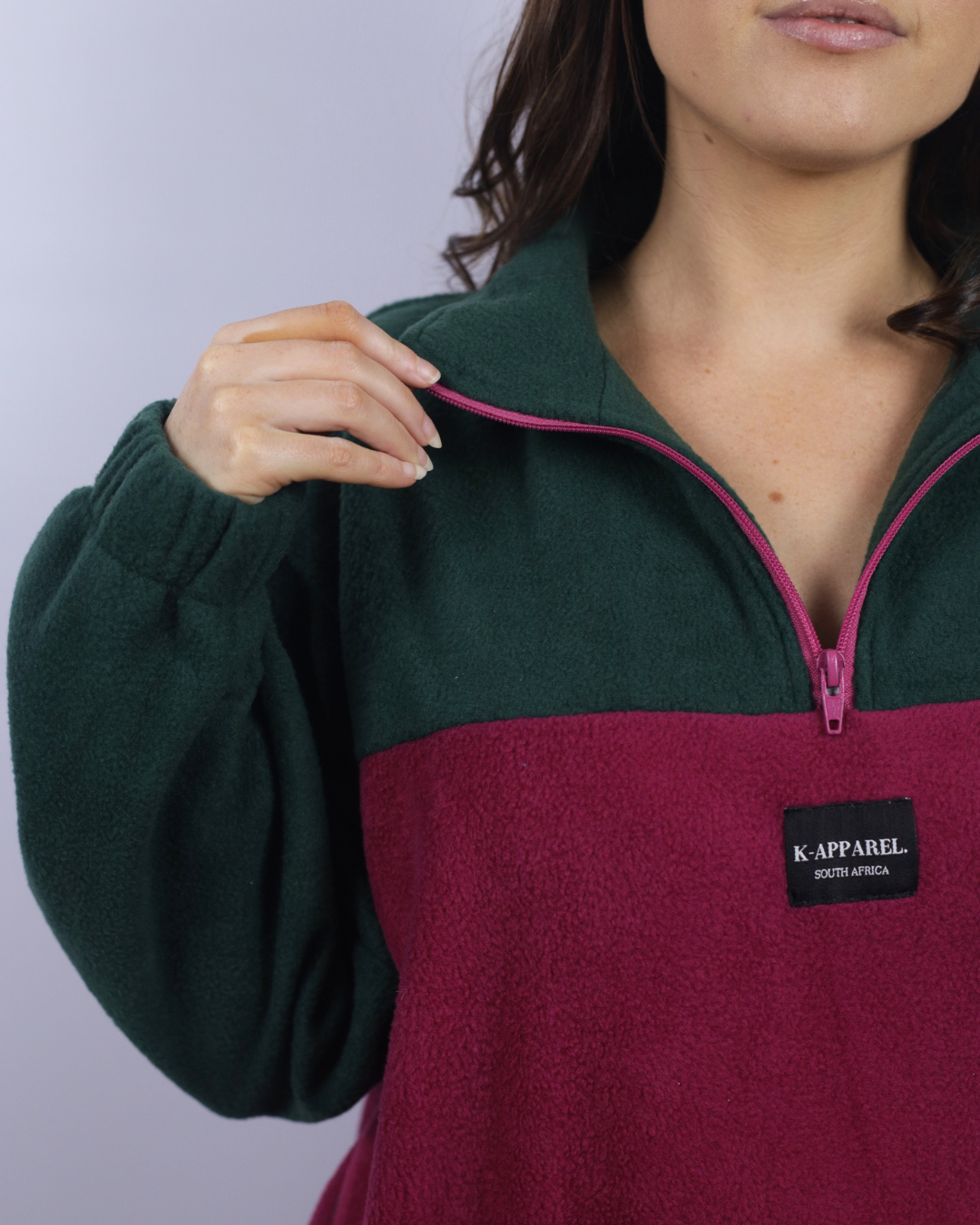 A close-up of a person wearing a color-block fleece pullover with a dark green upper section and a deep burgundy lower section, featuring a pink zipper and a black K-APPAREL logo patch.