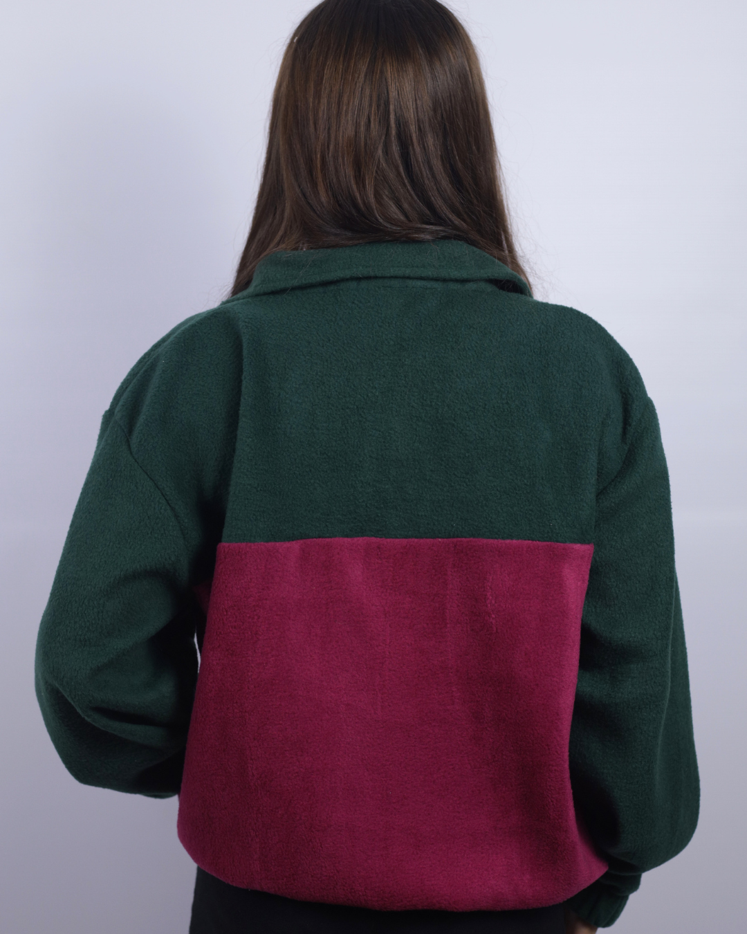 A person viewed from the back wearing a color-block fleece jacket with a dark green upper section and a deep magenta-pink lower back panel, set against a plain light gray background.