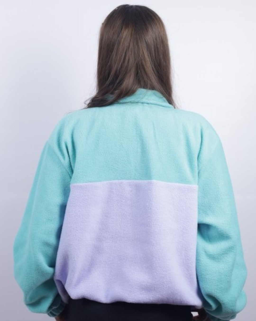 A person viewed from behind wearing a color-block fleece jacket with a mint green top section and a lavender purple bottom panel, set against a plain light gray background.