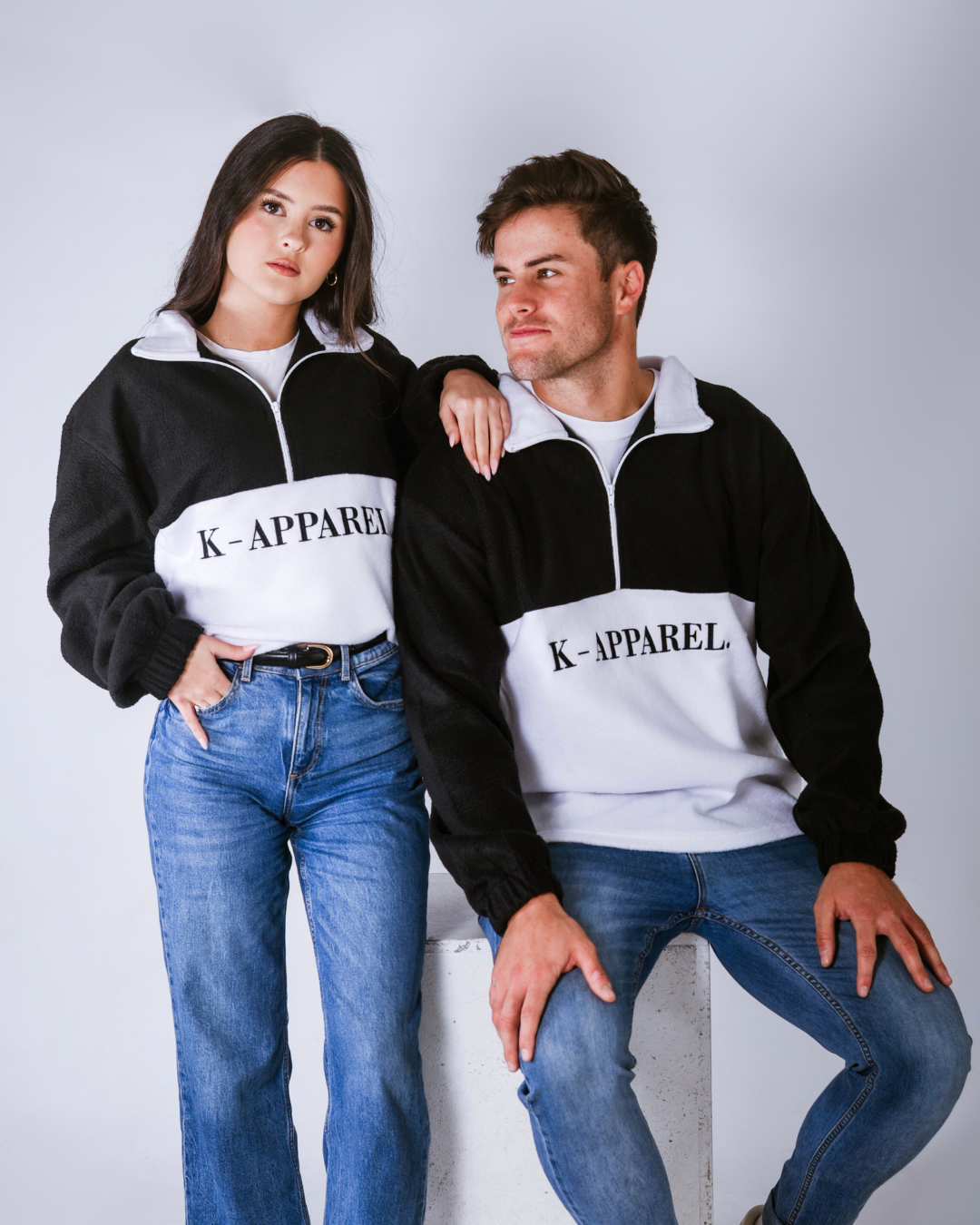 Two people wearing matching black and white  retro quarter zip fleece jerseys with visible branding on a plain background.