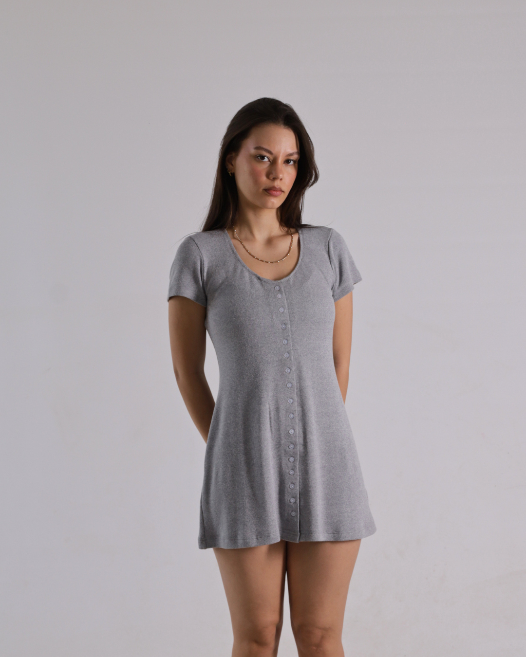 A woman modeling a light gray short-sleeve button-front tunic dress against a plain light gray studio background.