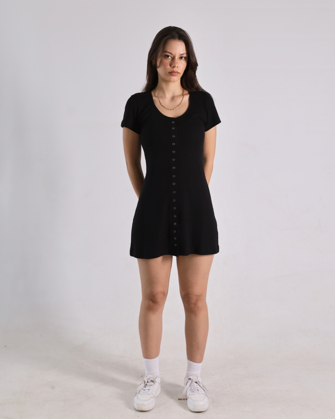 A woman modeling a black short-sleeve mini dress with a button-down front, paired with white socks and white sneakers, standing against a plain light gray studio background.