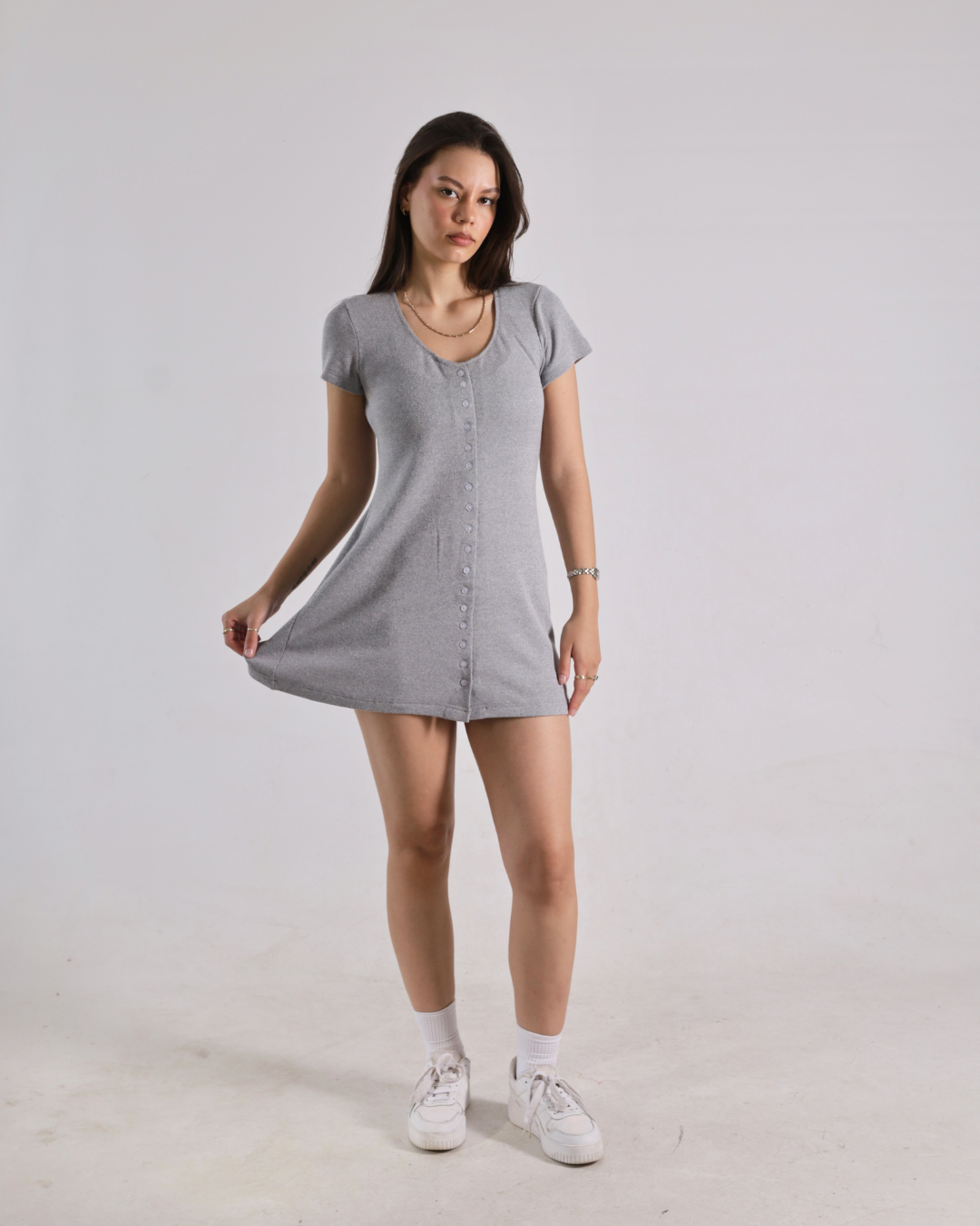 A woman modeling a light gray short-sleeve button-front mini dress with white socks and white sneakers, standing against a plain light gray studio background.