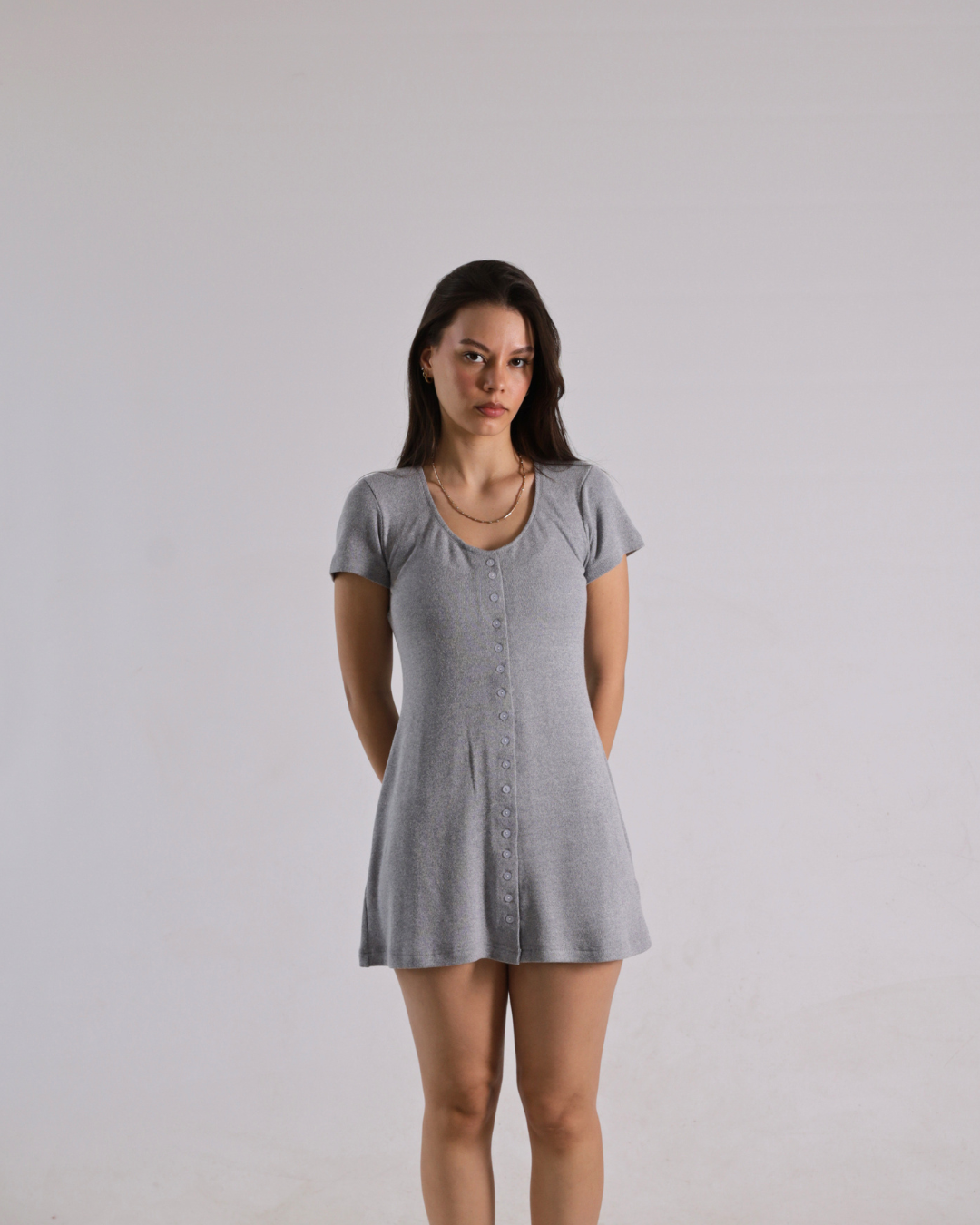 A woman modeling a short-sleeve gray mini dress with a button-up front, standing against a plain light gray studio background.