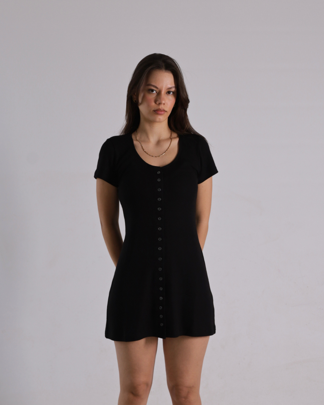 A woman modeling a short-sleeve black mini dress with a button-down front, standing against a plain light gray studio background.