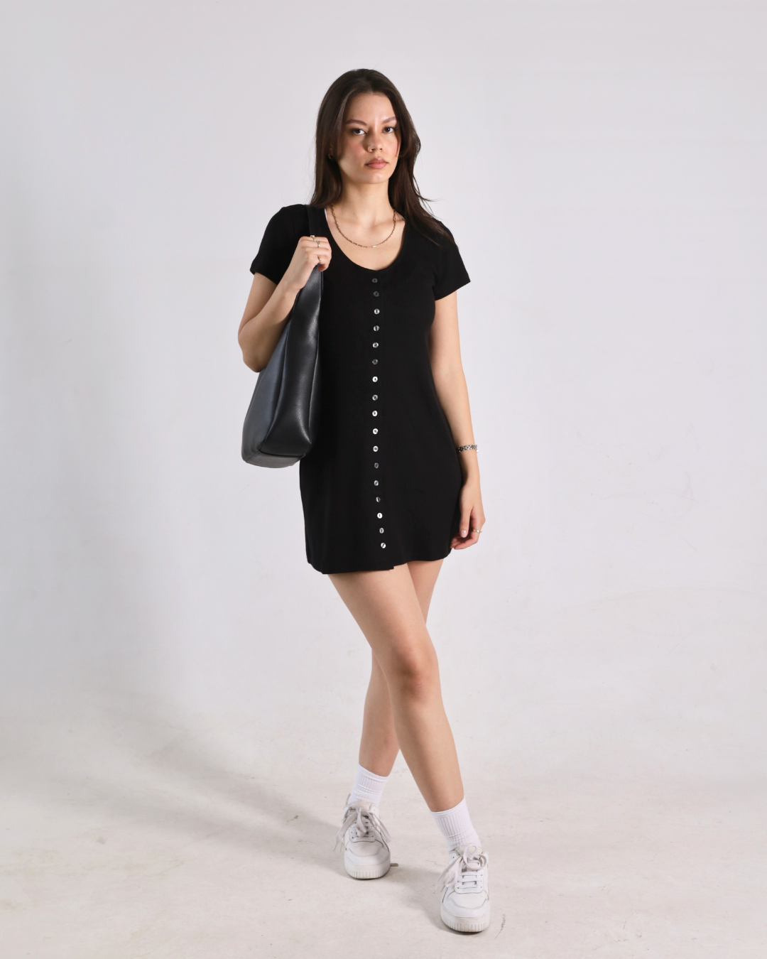 A woman modeling a black short-sleeve mini dress with a button-down front, styled with white socks, white sneakers, and a black shoulder bag against a plain light gray studio background.
