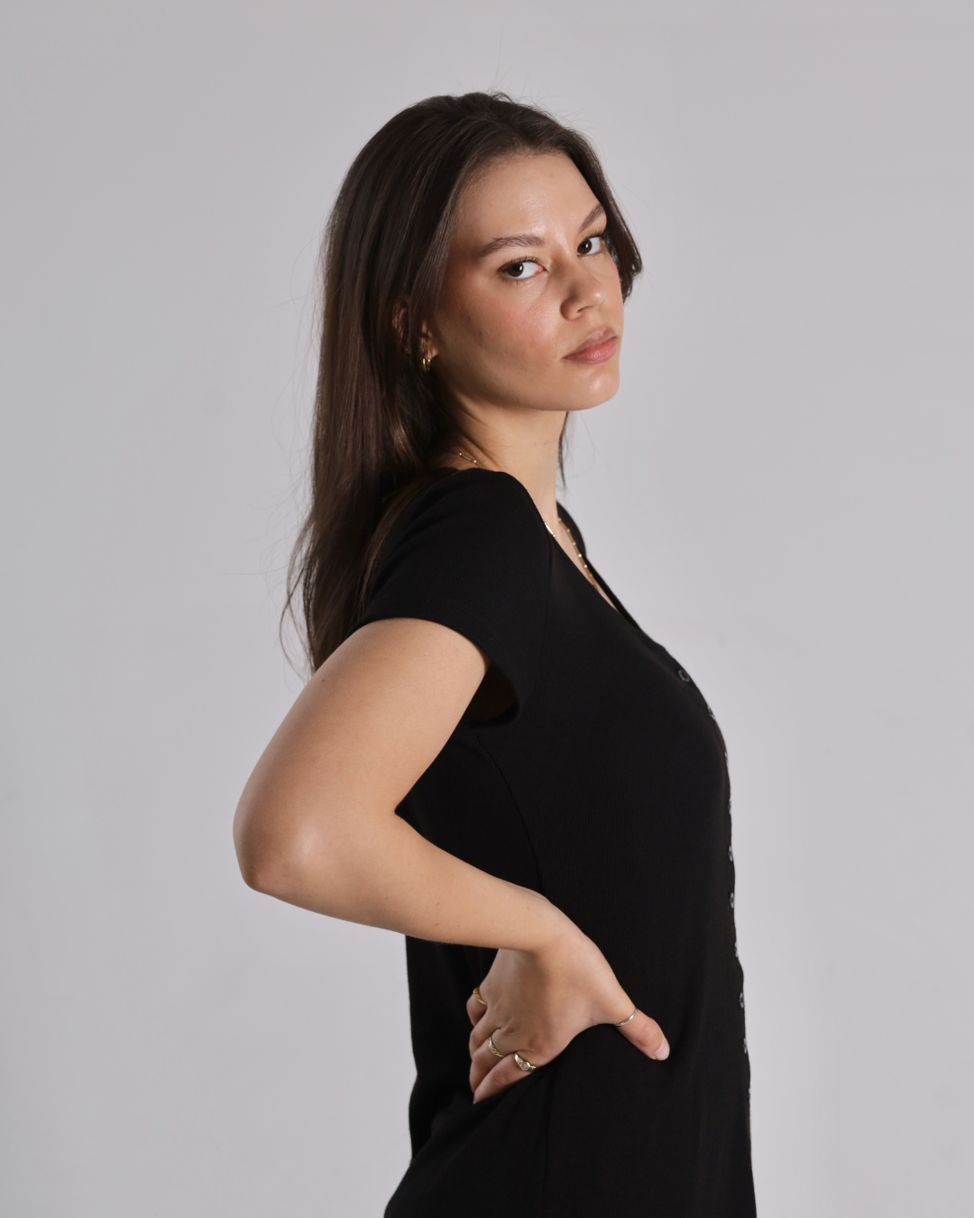 A woman with long dark hair is shown in profile wearing a black short-sleeve V-neck top against a plain light gray studio background.