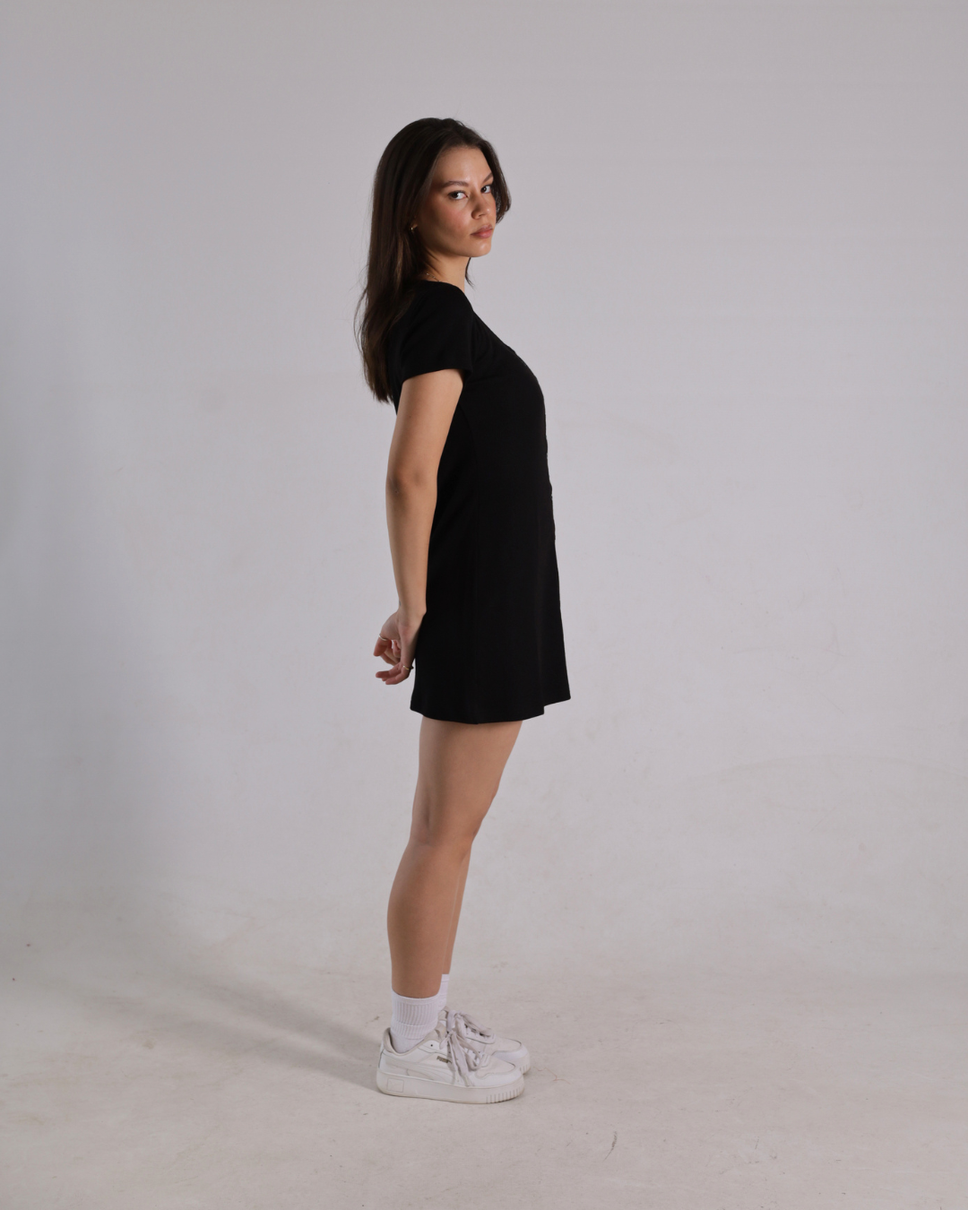 A woman stands in profile wearing a short black dress, white socks, and white sneakers against a plain light gray studio background.