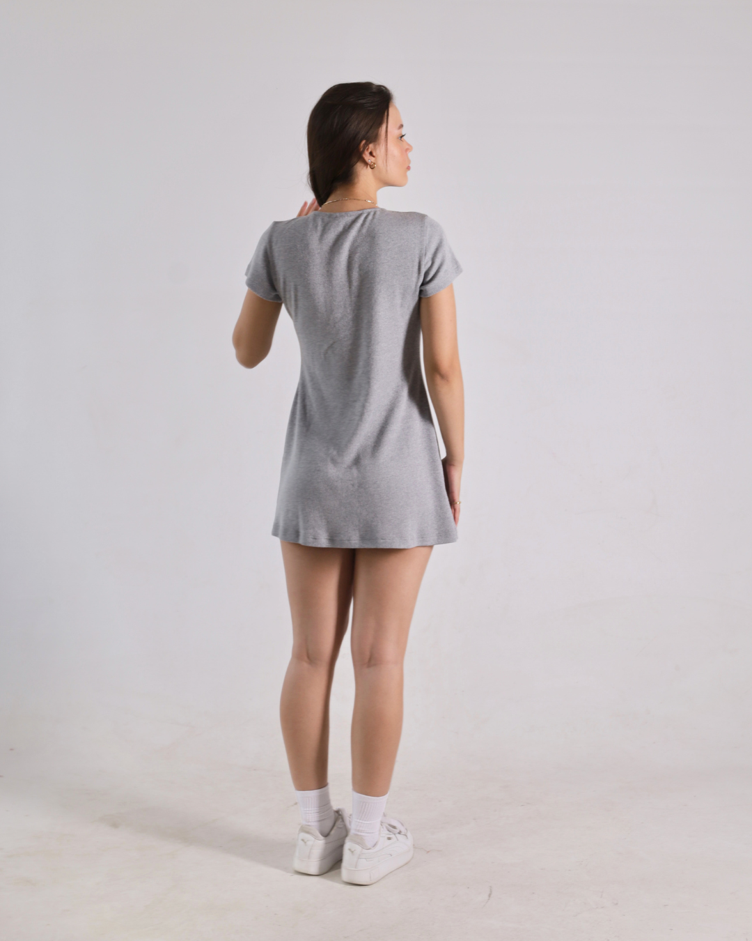 A woman is shown from behind wearing a short-sleeve gray t-shirt dress, white socks, and white sneakers against a plain light gray studio background.