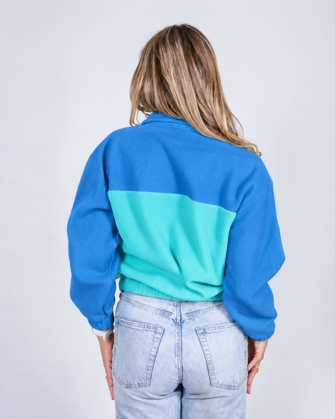 A woman viewed from behind wearing a color-blocked sweatshirt with a royal blue upper section and a turquoise lower section, paired with light-wash jeans against a plain light gray background.