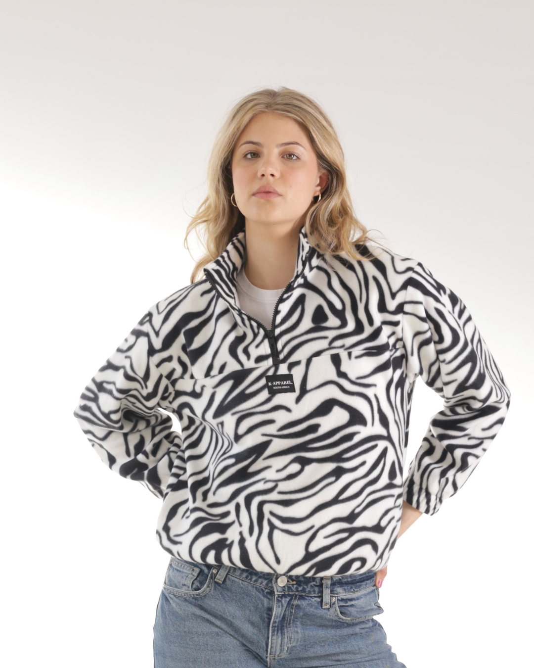 A woman modeling a black and white zebra-print fleece pullover jacket with a half-zip collar, paired with blue jeans, against a plain light gray studio background.