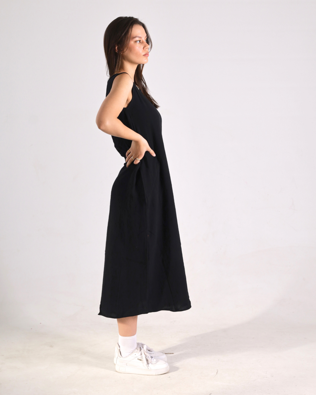 A woman modeling a sleeveless black midi dress with side pockets, paired with white socks and white sneakers, standing in profile against a plain white studio background.
