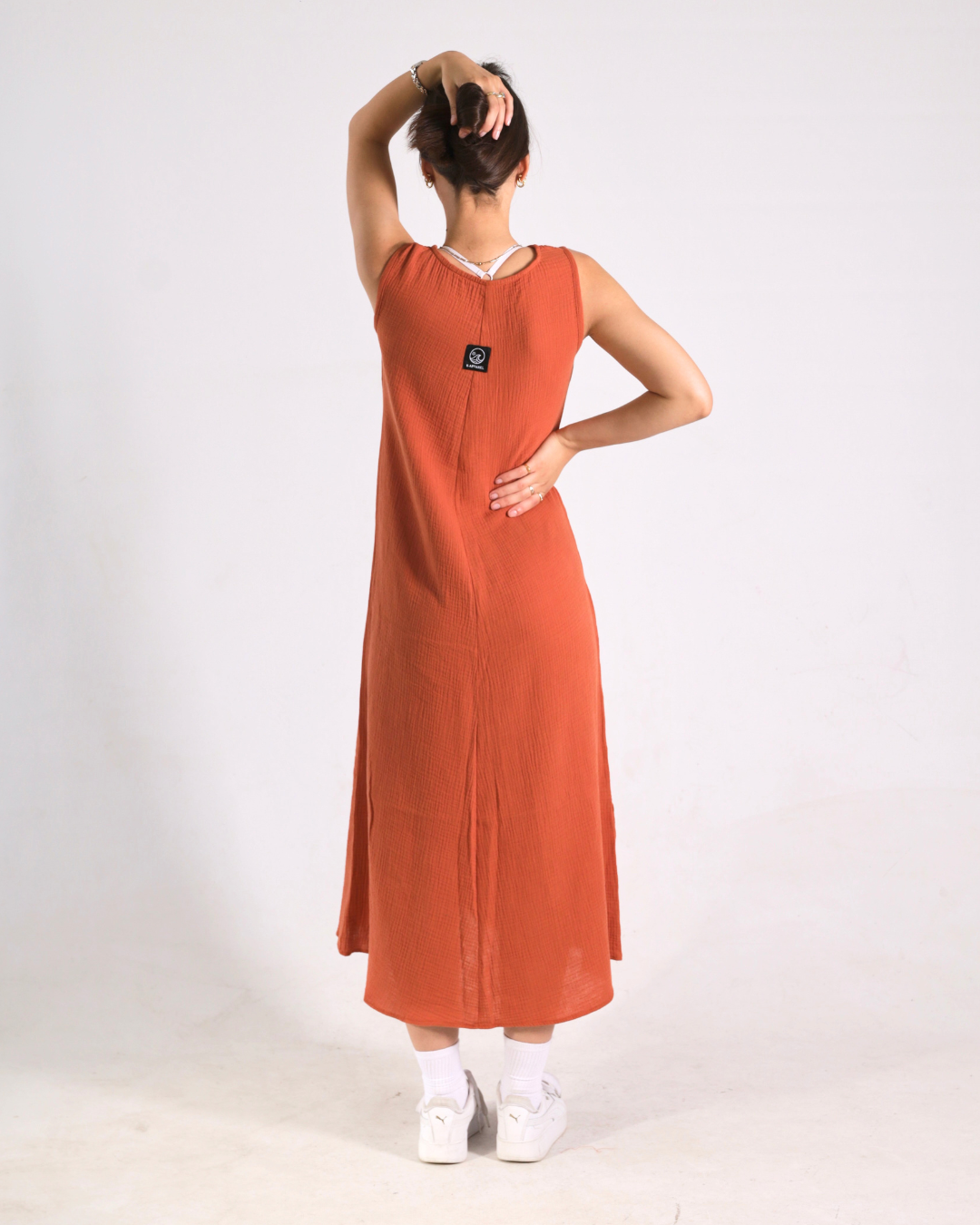 A woman wearing a sleeveless rust-orange maxi dress, viewed from the back. She has her hair in a bun and is wearing white socks and white sneakers against a plain white studio background.