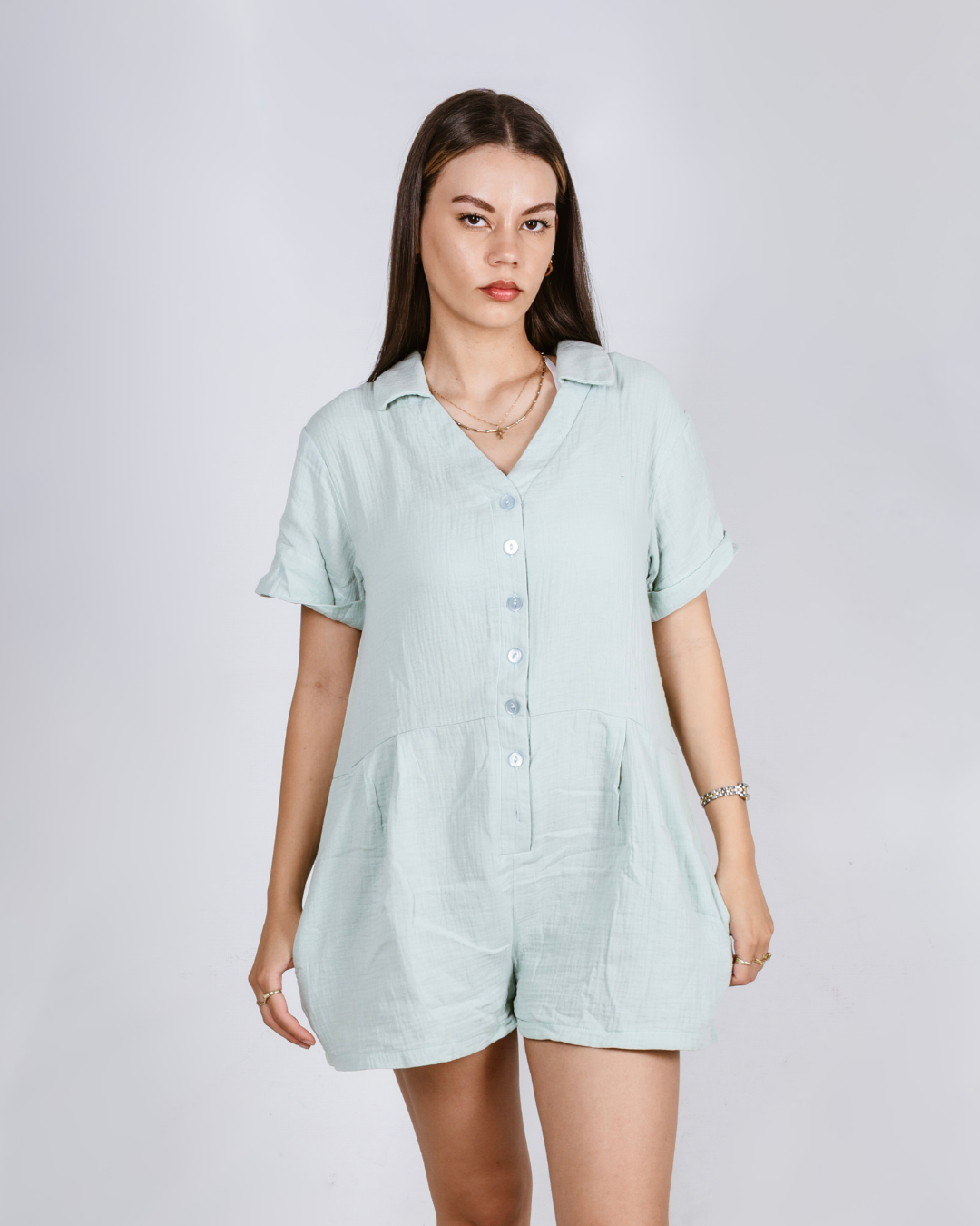 Woman wearing a sage button up cotton romper with pockets, against a white background