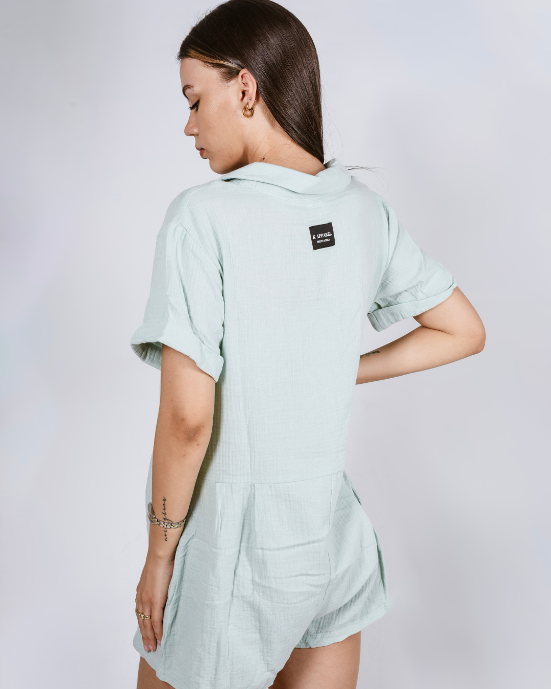 Woman wearing a sage button up cotton romper with pockets, against a white background